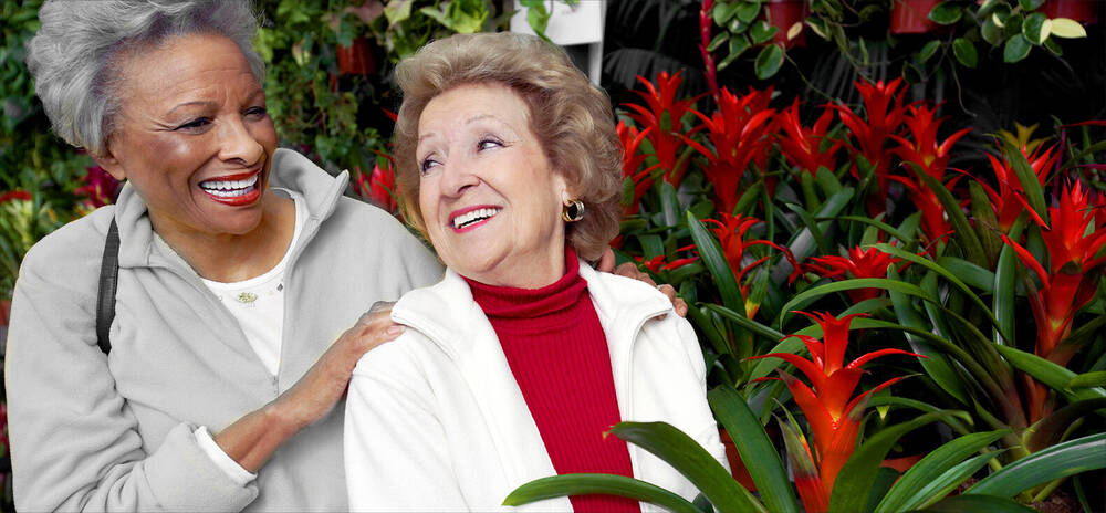 2 Elderly Women in front of red and green flowers.