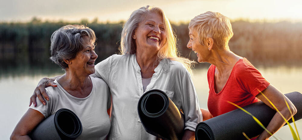 3 Elderly Women holding rolled yoga mats in front of a lake laughing together.